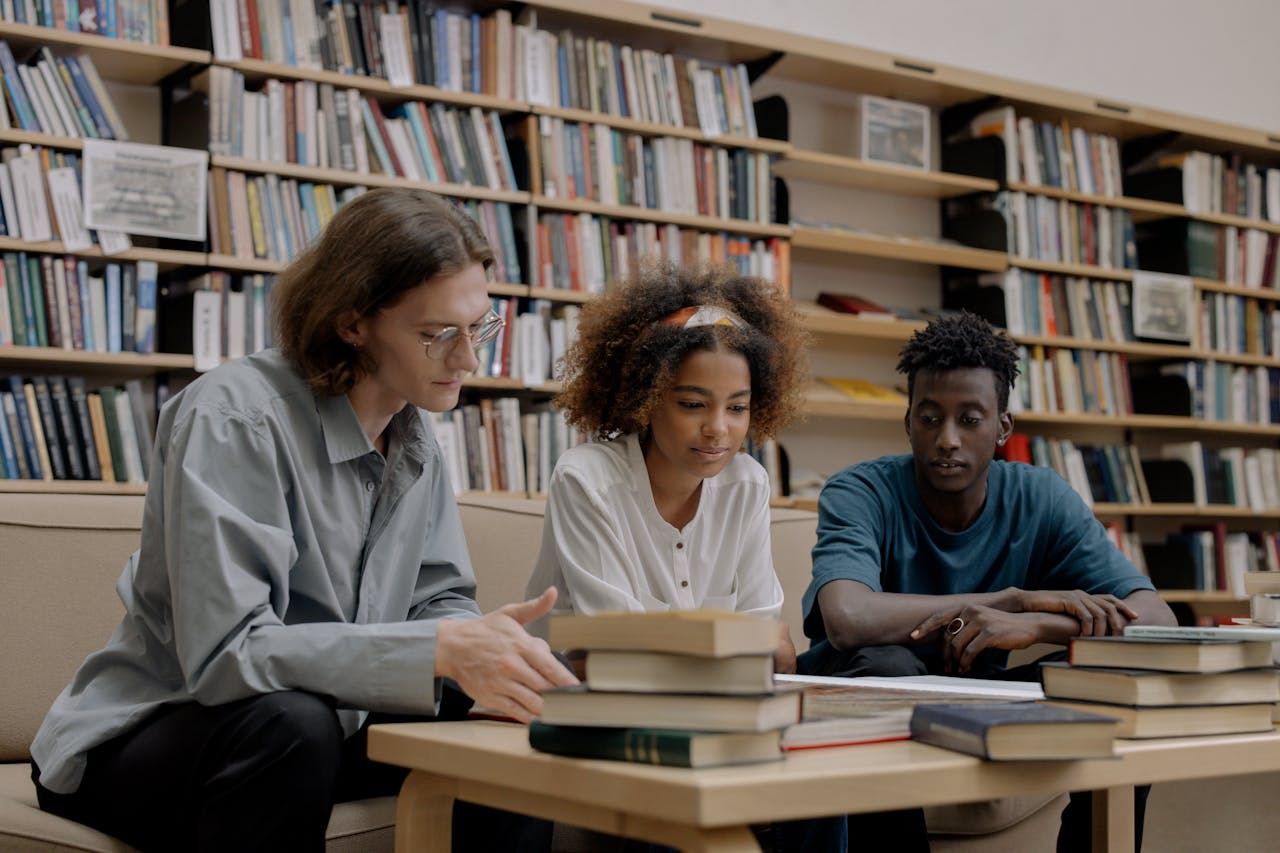 gallery-5 Three students studying together in a library, surrounded by books and learning materials.