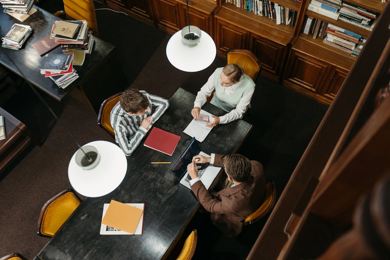 gallery-6 Three adults engaged in study at a library table, collaborating on academic work.